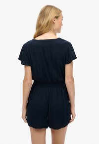Marineblå jumpsuit med korteærmer og elastisk talje. Har en scalloperet kant og blødt, let stof. Bagview vist.