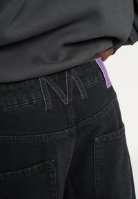 Person iført sorte denimjeans med et stort broderet "M" på taljen og et lilla mærke på højre bagside.