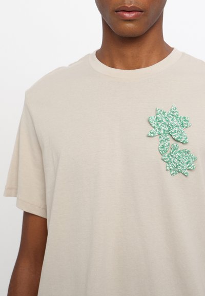 Beige cotton t-shirt featuring a green beaded floral appliqué on the left chest, round neckline, and short sleeves.