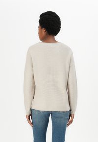 Pullover de malha bege com um decote largo e mangas longas, apresentando um acabamento texturizado e um rótulo discreto na parte de trás. Usado com jeans azuis.