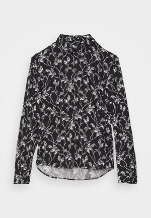 Personne portant une chemise noire à manches longues à fleurs, un pantalon blanc large et court, des chaussettes blanches et des chaussures blanches à lacets avec semelles en bois.