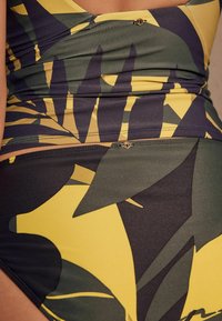 Bademode mit einem blättrigen Design in Grün und Gelb. Besitzt ein enganliegendes Oberteil und einen hoch taillierten Unterteil, hergestellt aus glattem, dehnbarem Stoff.