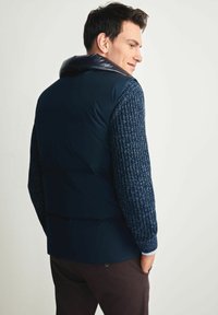 Granatowa kamizelka puchowa z wysokim kołnierzem i teksturowanymi długimi rękawami. Materiał jest gładki i puszysty, w kontraście do ściągaczowych rękawów.