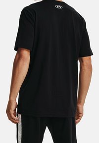 Hombre que lleva una camiseta negra de manga corta sin estampado con un pequeño logo blanco de Under Armour en la parte superior de la espalda.