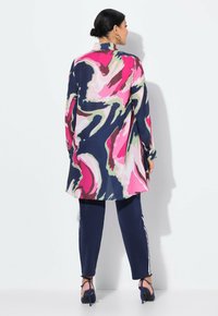 Blouse longue à motif abstrait bleu marine, rose et vert, associée à un pantalon bleu marine longueur cheville avec des rayures blanches sur les côtés. Sandales à talons.