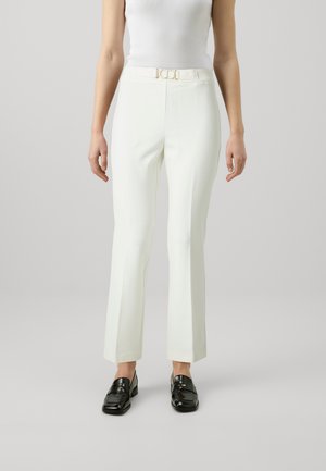 Pantaloni bianchi su misura con design a gamba svasata, dotati di una fibbia in tonalità oro. Indossati con un top in cotone aderente bianco e mocassini neri.