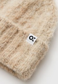 Cappello beanie lavorato a maglia beige con una texture morbida e pelosa. Presenta un risvolto e una piccola etichetta bianca con accenti neri del logo.