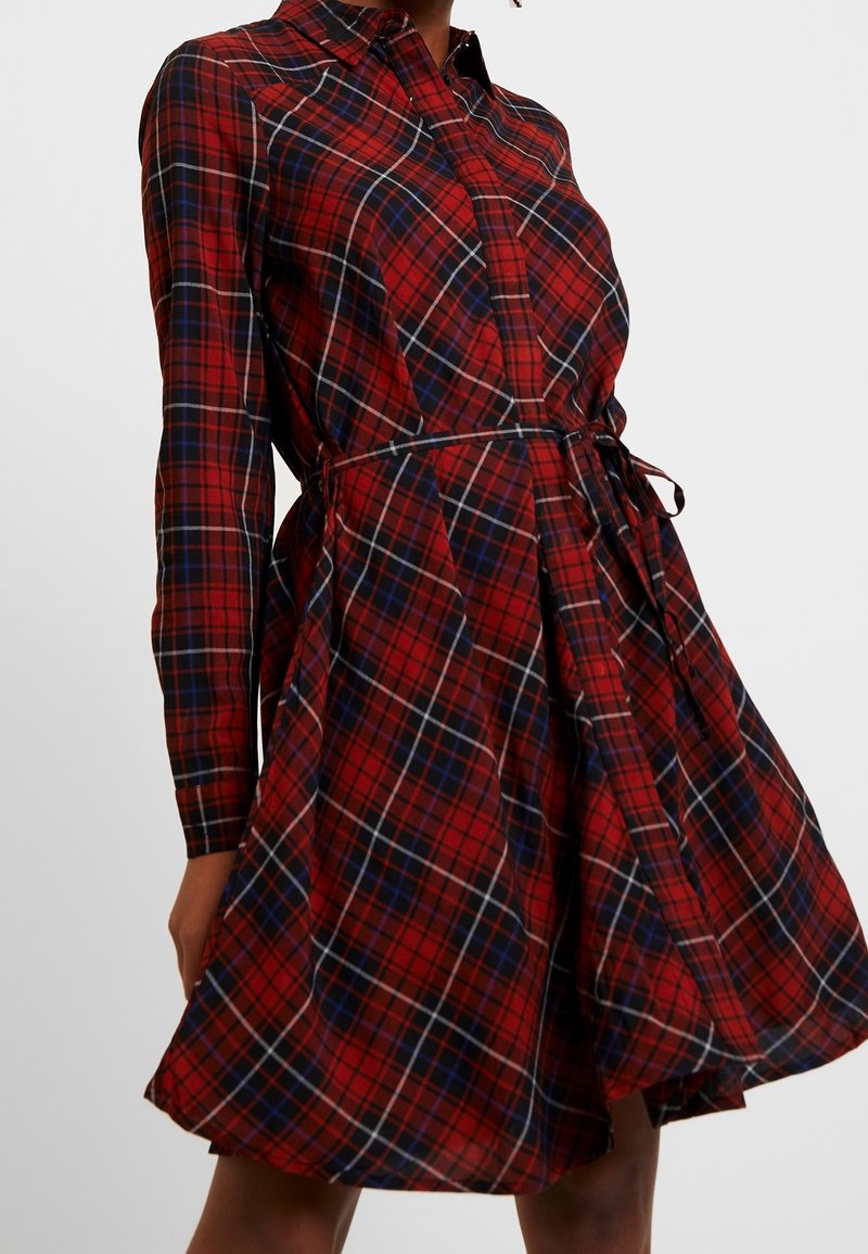 Punainen ja sininen ruudullinen mekko, jossa on pitkät hihat, kaulus ja vyö. Levittäytyvä hame kevyellä tekstuurilla ja napitus edessä.