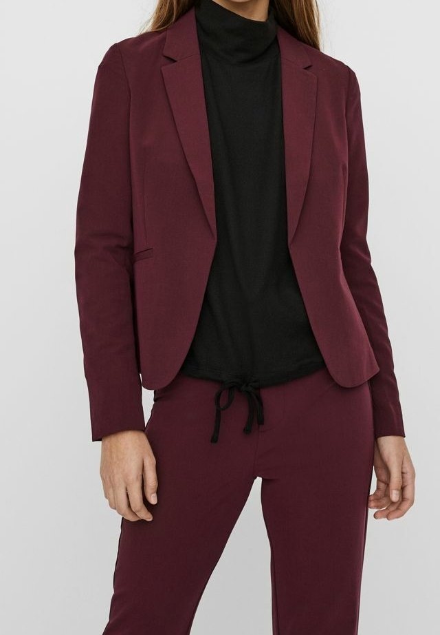 Femme portant un blazer ajusté bordeaux foncé et un pantalon assorti avec un haut noir à col montant en dessous.
