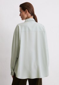 Woman with brown hair in a ponytail wearing an oversized pale green shirt viewed from the back against a plain white wall.