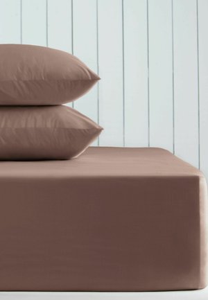 Zwei braune Kissen, auf einem passenden braunen Bett gestapelt, vor einem weißen Wandpaneel-Hintergrund.