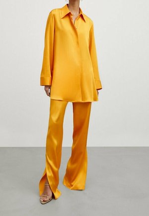 Femme portant une chemise en soie jaune doré brillante à manches longues et un pantalon large assorti avec des fentes avant, debout sur un sol gris.