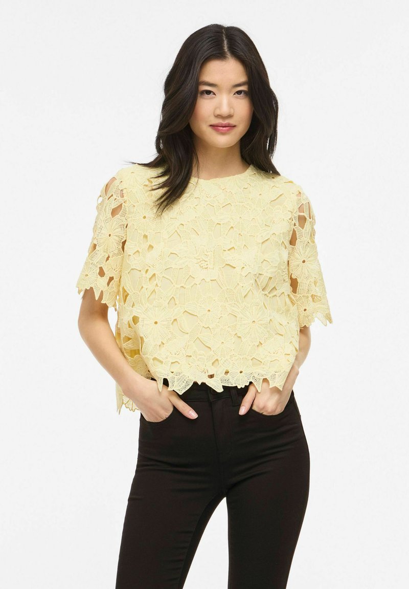 Jeune femme aux cheveux foncés portant un haut à manches courtes en dentelle fleurie jaune et un pantalon noir, debout les mains dans les poches.