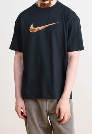 Schwarzes Baumwoll-T-Shirt mit einem großen, auffälligen braunen und orangefarbenen Nike-Swoosh-Logo auf der Brust. Kurze Ärmel, lockere Passform.