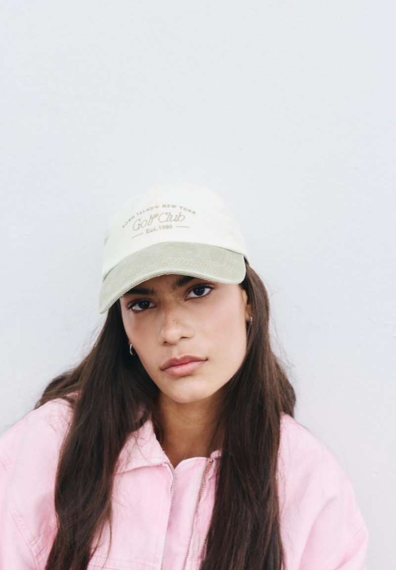 Jeune femme aux longs cheveux foncés portant une veste rose pâle et une casquette de baseball beige avec le texte "Golf Club" sur un fond blanc uni.