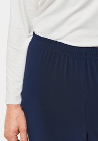 Navyblauwe broek met elastische tailleband en een soepele textuur, gecombineerd met een wit hemd met lange mouwen. Focus op taille- en stofdetails.