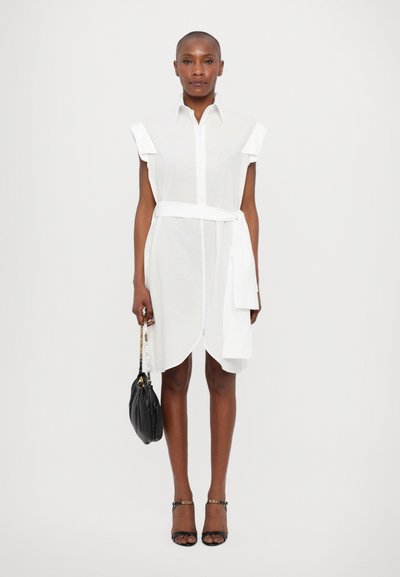 White shirt dress with short sleeves, belted waist, and curved hem. Paired with a black handbag and black strappy sandals.