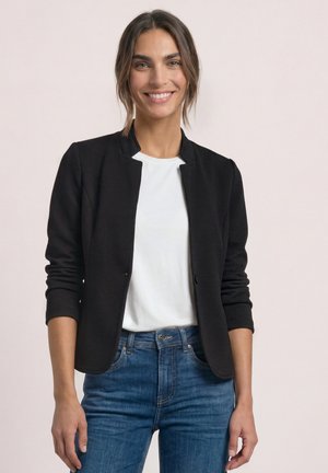 Femme souriante aux cheveux bruns portant un blazer noir texturé sur une chemise blanche et un jean bleu sur un fond clair uni.