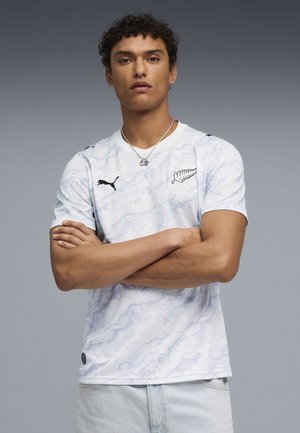 Joven con cabello rizado, brazos cruzados, usando una camiseta deportiva blanca con estampado de Puma con un logo de helecho plateado y jeans azul claro.