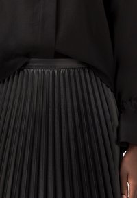 Jupe plissée en cuir noir avec une surface texturée, taille haute et ceinture lisse. Accompagnée d'un haut noir ample.