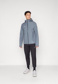 Grau wasserfeste Jacke mit Kapuze, Front-Reißverschluss, zwei Seitentaschen; kombiniert mit schwarzen Hosen und hellen Sneakern.