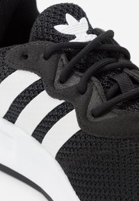 Svart träningssko med ett texturerat meshöverdrag, tre vita ränder, en gummisula och svarta skosnören. Har en vit logotyp på tungan.