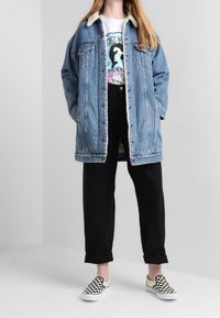 Blå denimjakke med shearling-krave, hvid T-shirt med grafik, sorte bukser med høj talje og sort-hvide ternede slip-on sko.