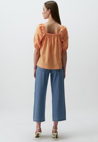 Blouse à manches bouffantes couleur pêche avec un décolleté carré, associée à un jean en denim bleu clair à jambes larges et des talons légers. Texture lisse, coupe décontractée.