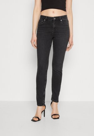 Vrouw draagt zwarte skinny jeans en zwarte sandaal met hoge hakken, een cropped zwart topje dat de buik laat zien, staand op een witte vloer tegen een witte muur.