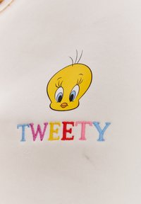 Bílá látka s žlutou hlavou karikaturního ptáčka s modrýma očima a černými akcenty, vedle slova "TWEETY" v vícebarevné výšivce.