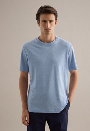 Jonge man met bruin haar, gekleed in een lichtblauw overhemd met korte mouwen en een donkere broek, staand tegen een effen beige achtergrond.