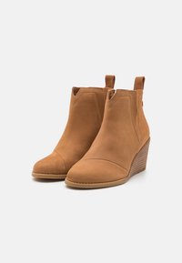 Bottines chevilles en daim beige avec un bout arrondi, dotées d'un talon compensé et de deux languettes à la base pour un enfilage facile.