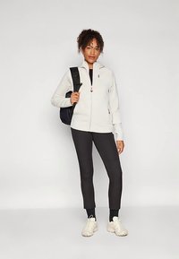 Casaco branco de tecido polar com fecho de correr e gola alta, leggings pretas e ténis de cor clara. A modelo leva uma mochila preta.