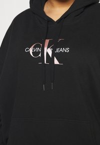 Czarna bawełniana bluza z kapturem, z przednią kieszenią, kapturem ze sznurkiem oraz dużym nadrukiem logo w metalicznym różowym złocie z napisem "CALVIN KLEIN JEANS."