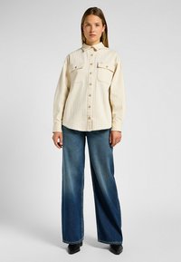 Camicia button-up color crema con righe verticali, due tasche sul petto e colletto, abbinata a jeans di denim blu a gamba larga.