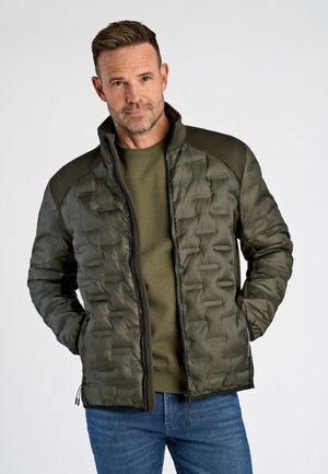Giacca oliva trapuntata con colletto alto, chiusura con zip e dettagli testurizzati sulle spalle. Abbinata a un maglione verde e jeans in denim.