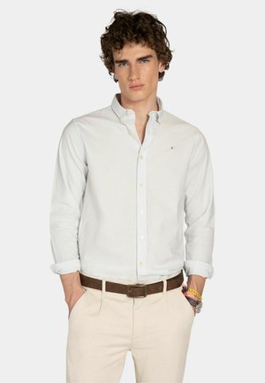 Joven con cabello rizado, viste una camisa de botones clara con rayas, pantalones beige, cinturón marrón y pulseras, de pie con las manos en los bolsillos.