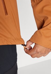 Brown softshell jacket with an adjustable drawstring hem. The sleeve features an orange accent and a snap closure. Smooth, textured fabric.