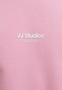 Roosa puuvillane dressipluus, millel on ümar kaelus ja tikitud valge logo "JJ Studios" rinnal. Sile tekstuur ja minimaalne detailitus.