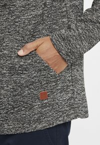 Suéter con una textura de tela gris jaspeado. Presenta una etiqueta de cuero marrón, un bolsillo frontal y un corte relajado con detalles de costura visibles.