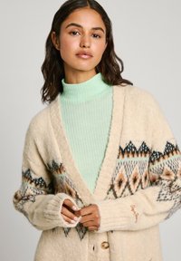 Cardigan crema con motivo geometrico in tonalità di marrone, nero e accenti di verde teal, dotato di chiusura con bottoni e polsini a coste sopra un dolcevita verde menta.
