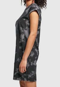 Personne portant une robe tie-dye noire et grise à manches courtes, longueur genou, debout de profil sur un fond uni.