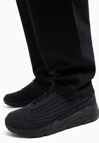 Scarpe da ginnastica nere con texture rigata, dotate di una suola spessa e di un design minimalista, accostate a pantaloni neri aderenti, parzialmente visibili.