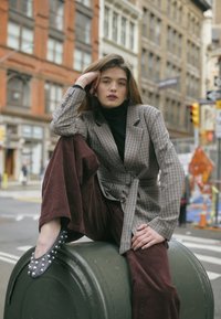 Kariertes Blazer mit tailliertem Gürtel in neutralen Tönen, kombiniert mit burgunderfarbenen Cordhosen und schwarzen, mit Nieten versehenen Ballerinas, sitzend auf einem grünen Fass.