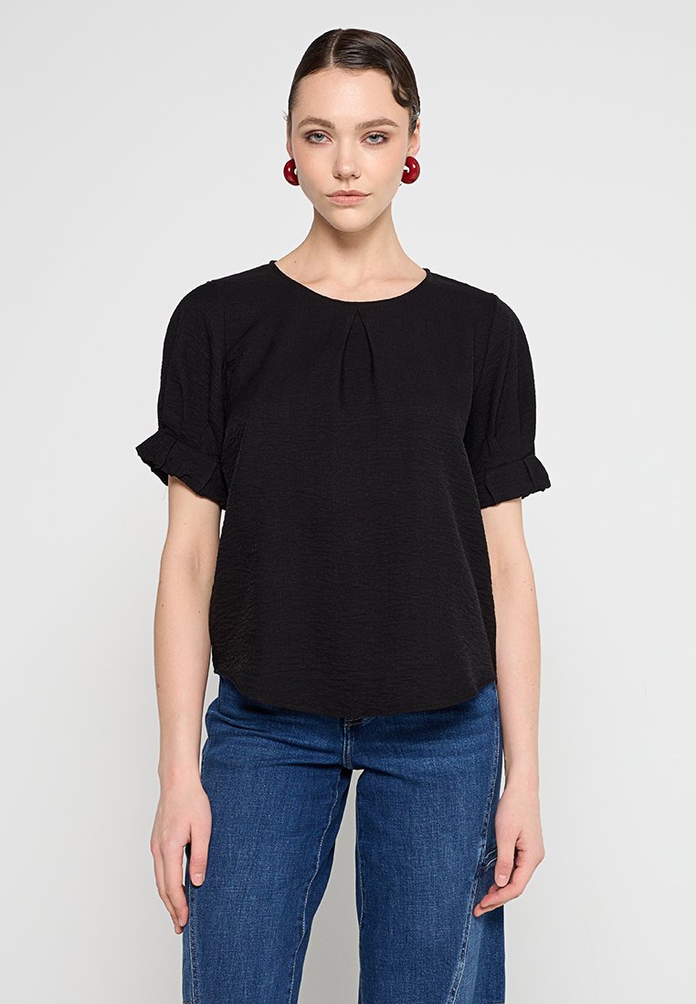 Schwarzes Kurzarmoberteil mit rundem Halsausschnitt und Faltendetails. Leichter, strukturierten Stoff mit fransigen Ärmelakzenten. Getragen mit blauer Jeans.