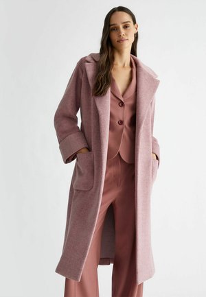 Cappotto lungo rosa a spina di pesce con ampio colletto, grandi tasche e polsini arrotolati, indossato sopra un completo rosa abbinato. Texture liscia.