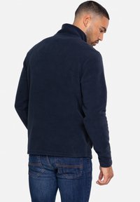 Marineblå fleecejakke med høj krave, lange ærmer og en blød tekstur. Kombineret med blå jeans med syede baglommer.