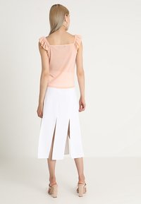 Top pêche avec des manches froncées et un tissu texturé ; assorti à une jupe blanche avec des fentes sur les côtés. Sandales à talons blocs neutres.