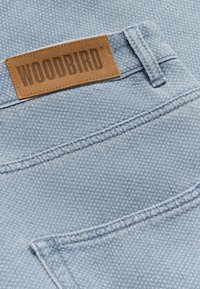 Tissu en denim bleu clair texturé avec couture de poche arrière et patch en cuir marron marqué WOODBIRD au-dessus de la passants de ceinture.