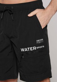 Fekete vízisport rövidnadrág könnyű anyagból, állítható húzózsinórral, oldalsó zsebekkel és feltűnő fehér felirattal: „VÍZISPORT.”
